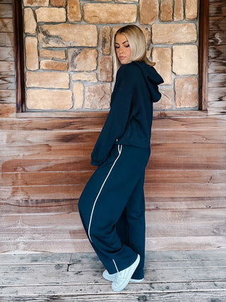 A person wearing teal navy wide-leg sweatpants with white side stripes, paired with a matching hoodie and white sneakers, standing against a rustic wooden wall.