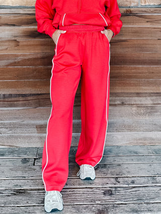 A person wearing bright red sweatpants with white side stripes and a matching red hoodie, standing against a rustic wooden plank wall.