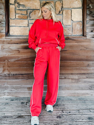 A person wearing bright red sweatpants with white side stripes and a matching red hoodie, standing against a rustic wooden plank wall.