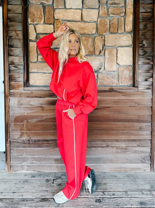 A person wearing bright red sweatpants with white side stripes and a matching red hoodie, standing against a rustic wooden plank wall.