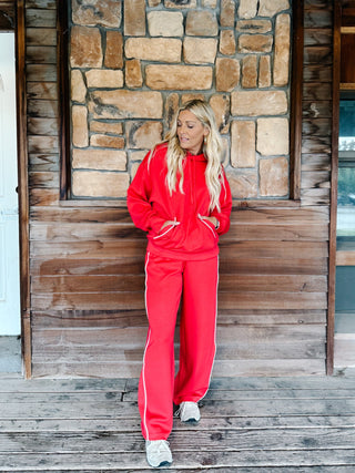 A person wearing bright red sweatpants with white side stripes and a matching red hoodie, standing against a rustic wooden plank wall.