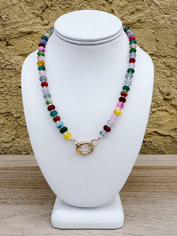 A colorful beaded necklace with a gold clasp displayed on a white jewelry bust against a textured tan wall and light wood surface.