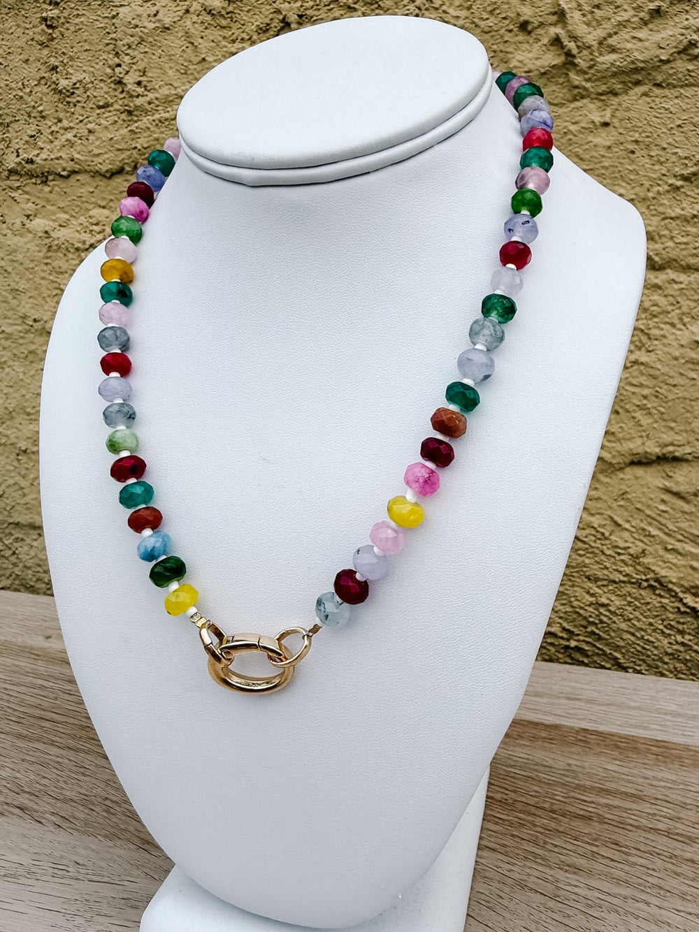 A colorful beaded necklace with a gold clasp displayed on a white mannequin bust against a textured tan wall.