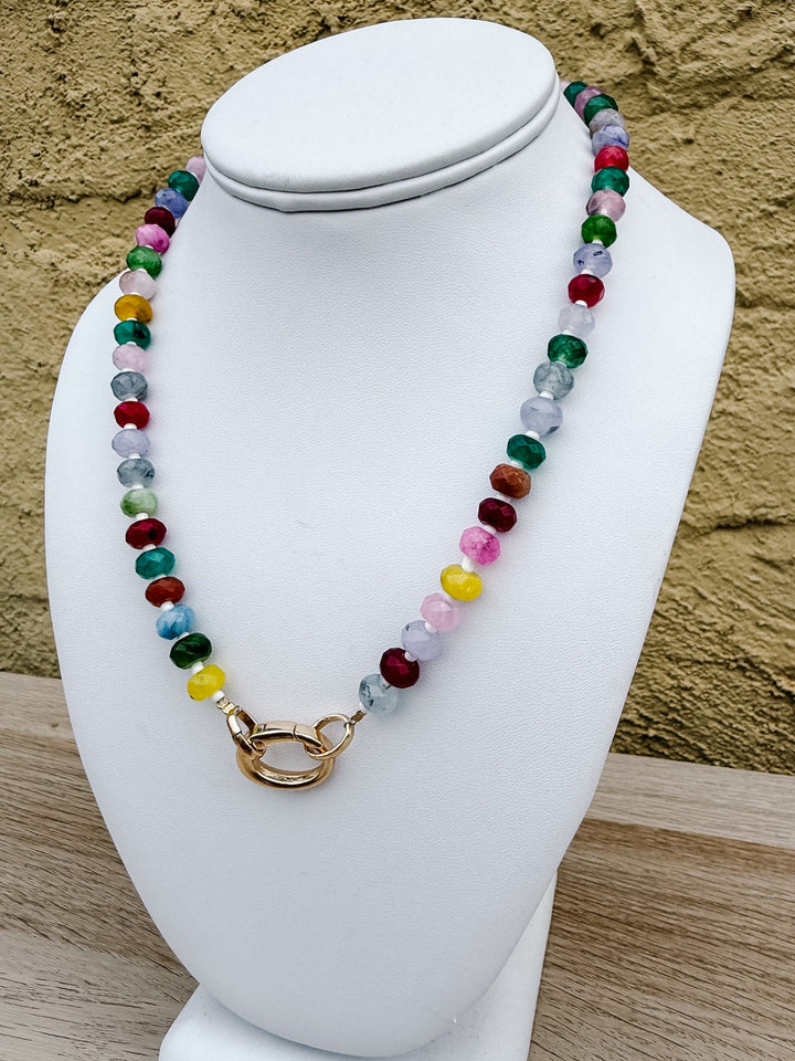 A colorful beaded necklace with a gold clasp displayed on a white mannequin bust against a textured tan wall.