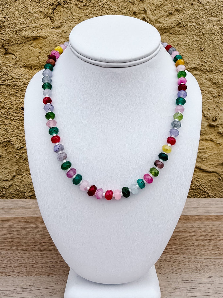 A multicolor beaded necklace made of polished round gemstone beads, displayed on a white ceramic jewelry bust against a textured tan stucco wall and light wood surface.