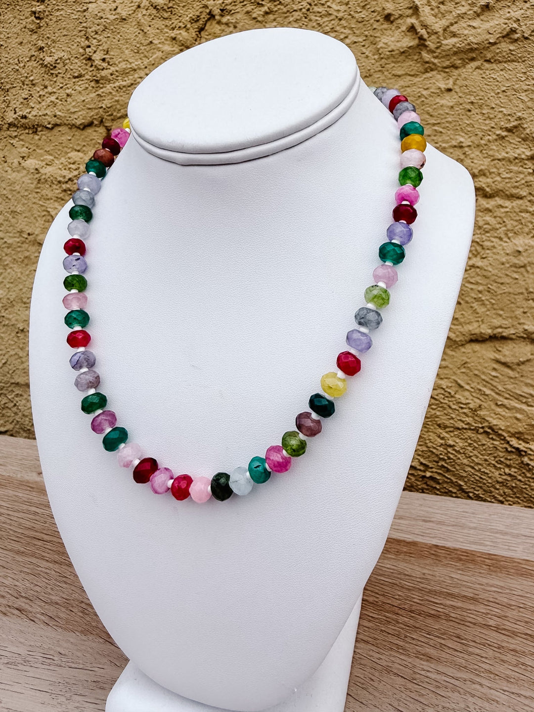 A multicolor beaded necklace made of polished round gemstone-like beads, displayed on a white jewelry bust against a textured tan wall and light wood surface.