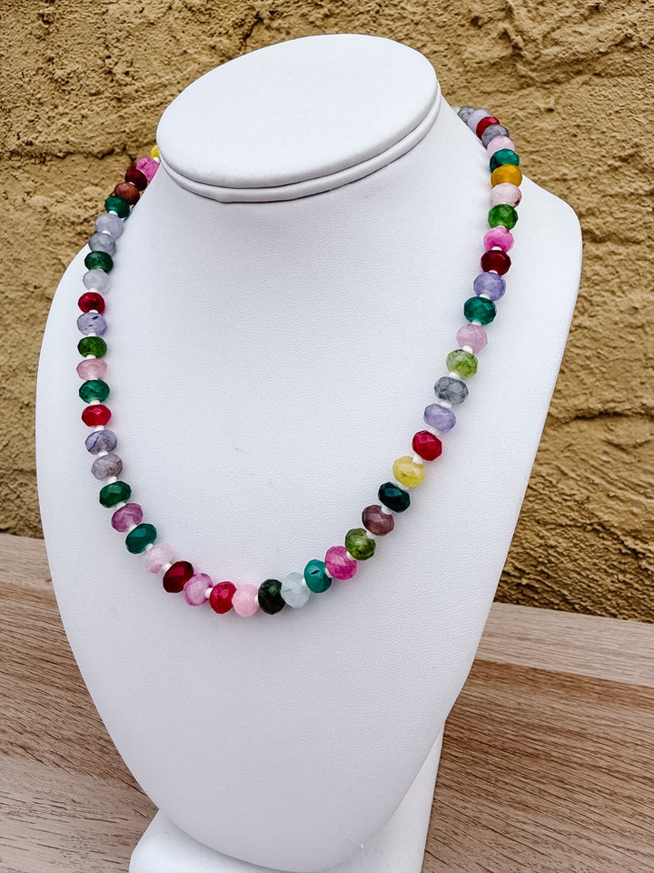 A multicolor beaded necklace made of polished round gemstone-like beads, displayed on a white jewelry bust against a textured tan wall and light wood surface.