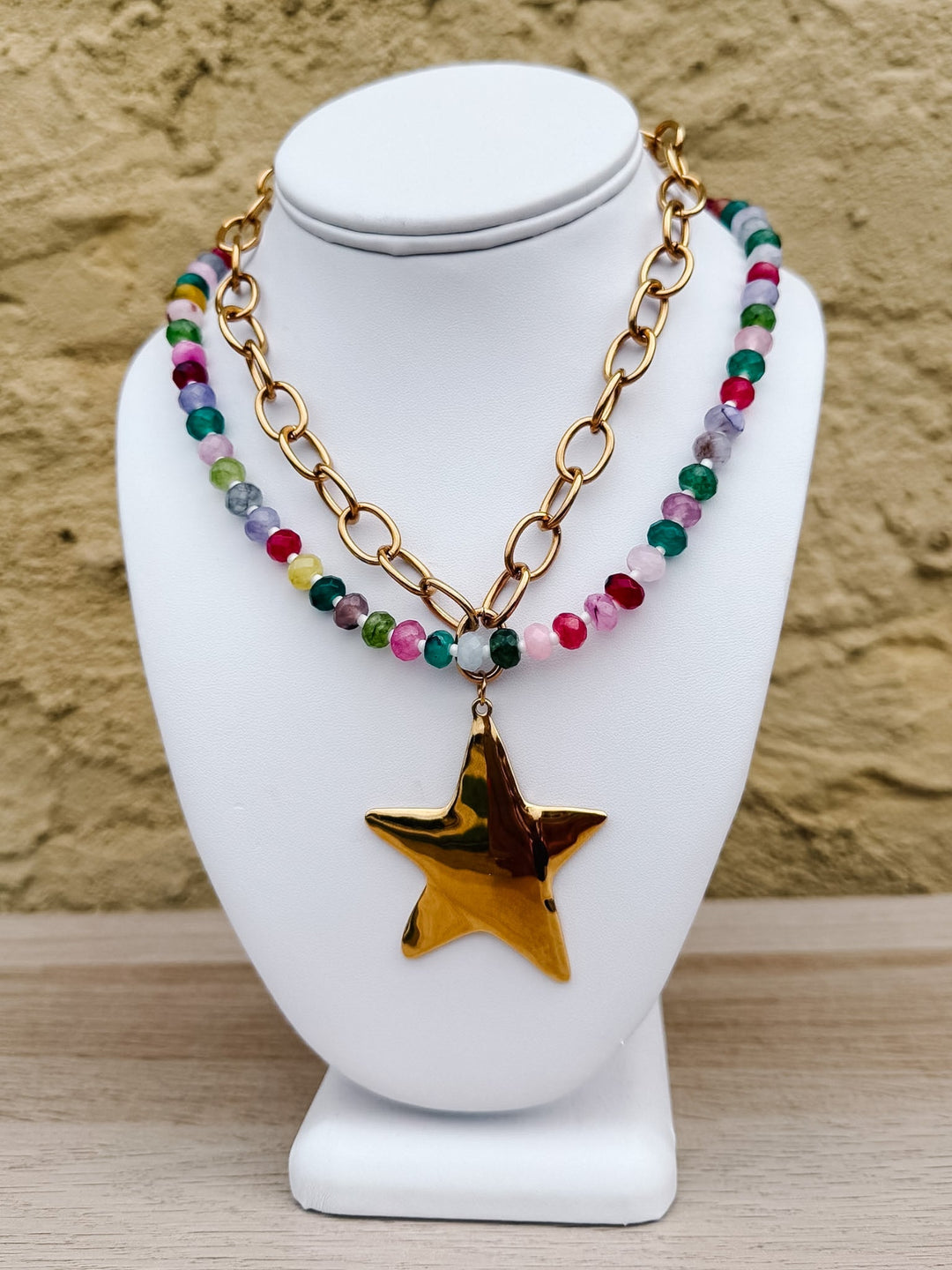 A double-strand necklace displayed on a white mannequin bust. One strand features a gold link chain, and the other is made of small multicolored beads with a dangling gold star pendant.
