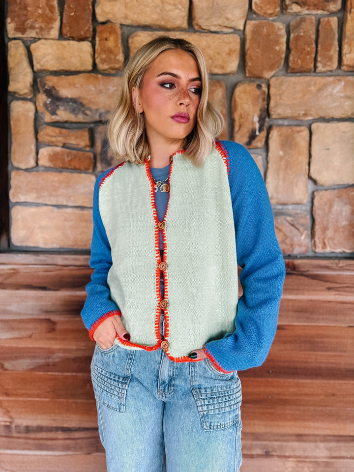 A woman wearing a blue knit sweater vest with a sage green front panel and orange-red trim, paired with light-wash jeans, standing in front of a rustic stone wall.