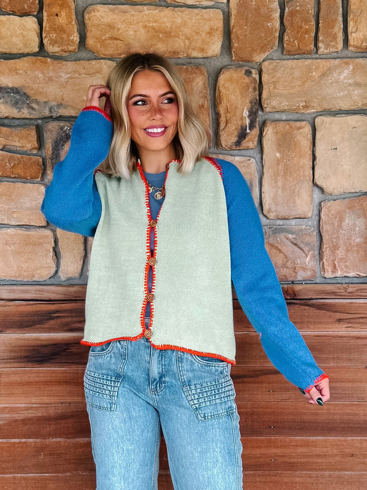 A woman wearing a sage green cardigan with blue sleeves and a orange trim over a blue tank and light blue jeans, standing in front of a rustic stone wall.