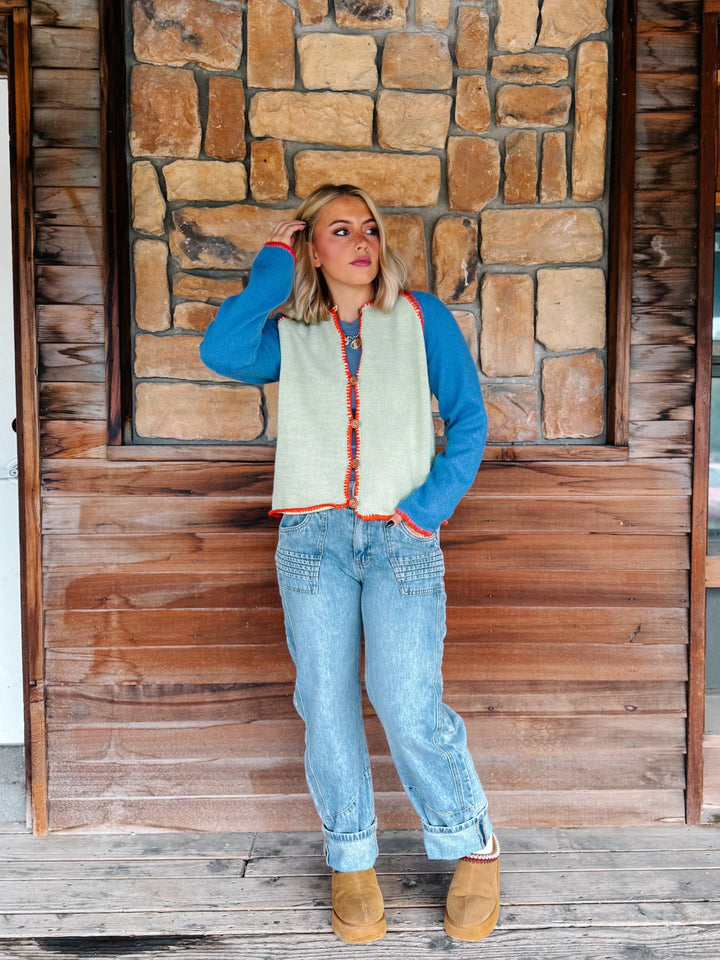 A woman wearing a sage green cardigan with blue sleeves and a orange trim over a blue tank and light blue jeans, standing in front of a rustic stone wall.