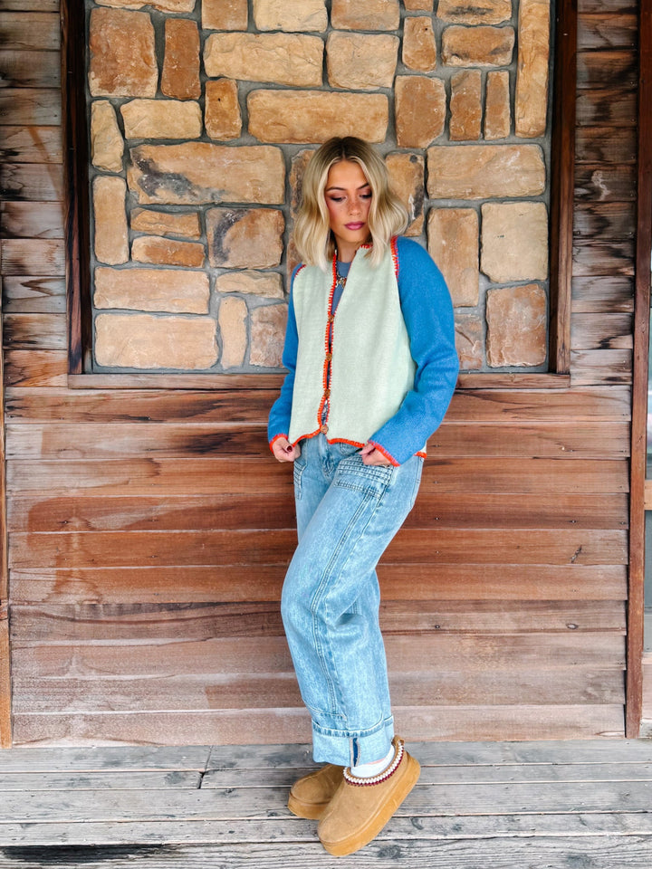 A woman wearing a sage green cardigan with blue sleeves and a orange trim over a blue tank and light blue jeans, standing in front of a rustic stone wall.