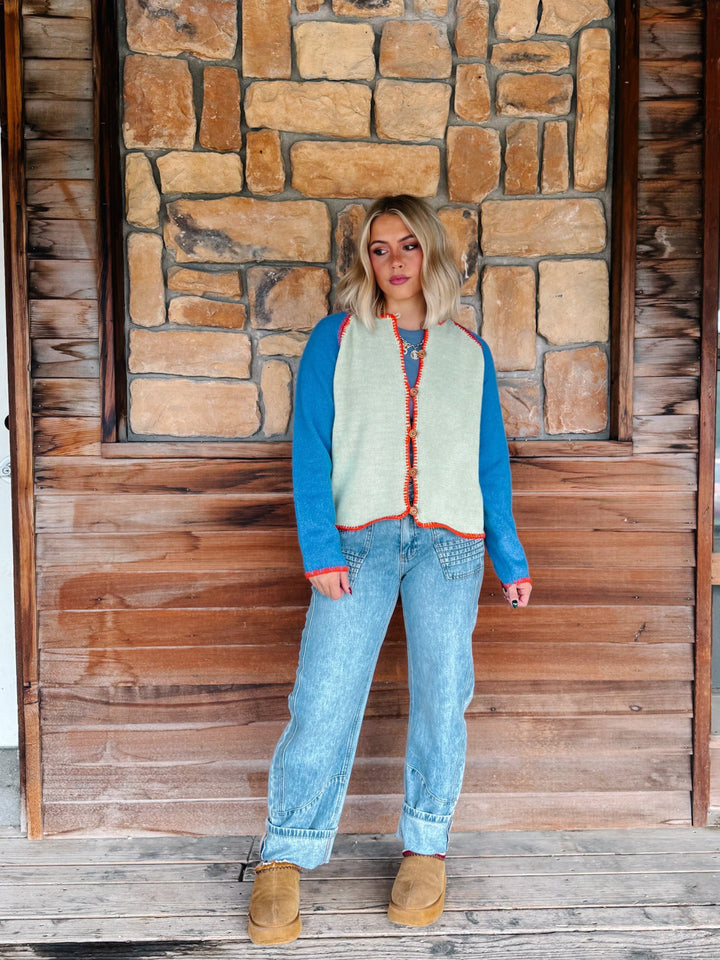 A woman wearing a sage green cardigan with blue sleeves and a orange trim over a blue tank and light blue jeans, standing in front of a rustic stone wall.