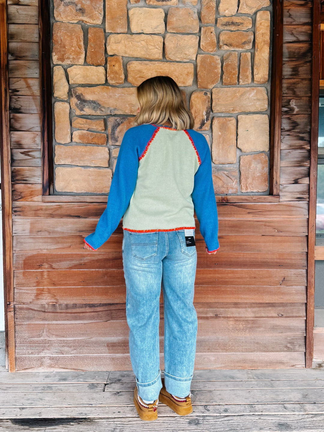 A woman wearing a sage green cardigan with blue sleeves and a orange trim over a blue tank and light blue jeans, standing in front of a rustic stone wall.