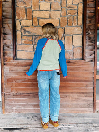 A woman wearing a sage green cardigan with blue sleeves and a orange trim over a blue tank and light blue jeans, standing in front of a rustic stone wall.