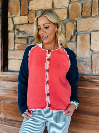A woman wearing a coral pink and navy blue raglan cardigan with brown buttons, standing in front of a rustic stone wall.