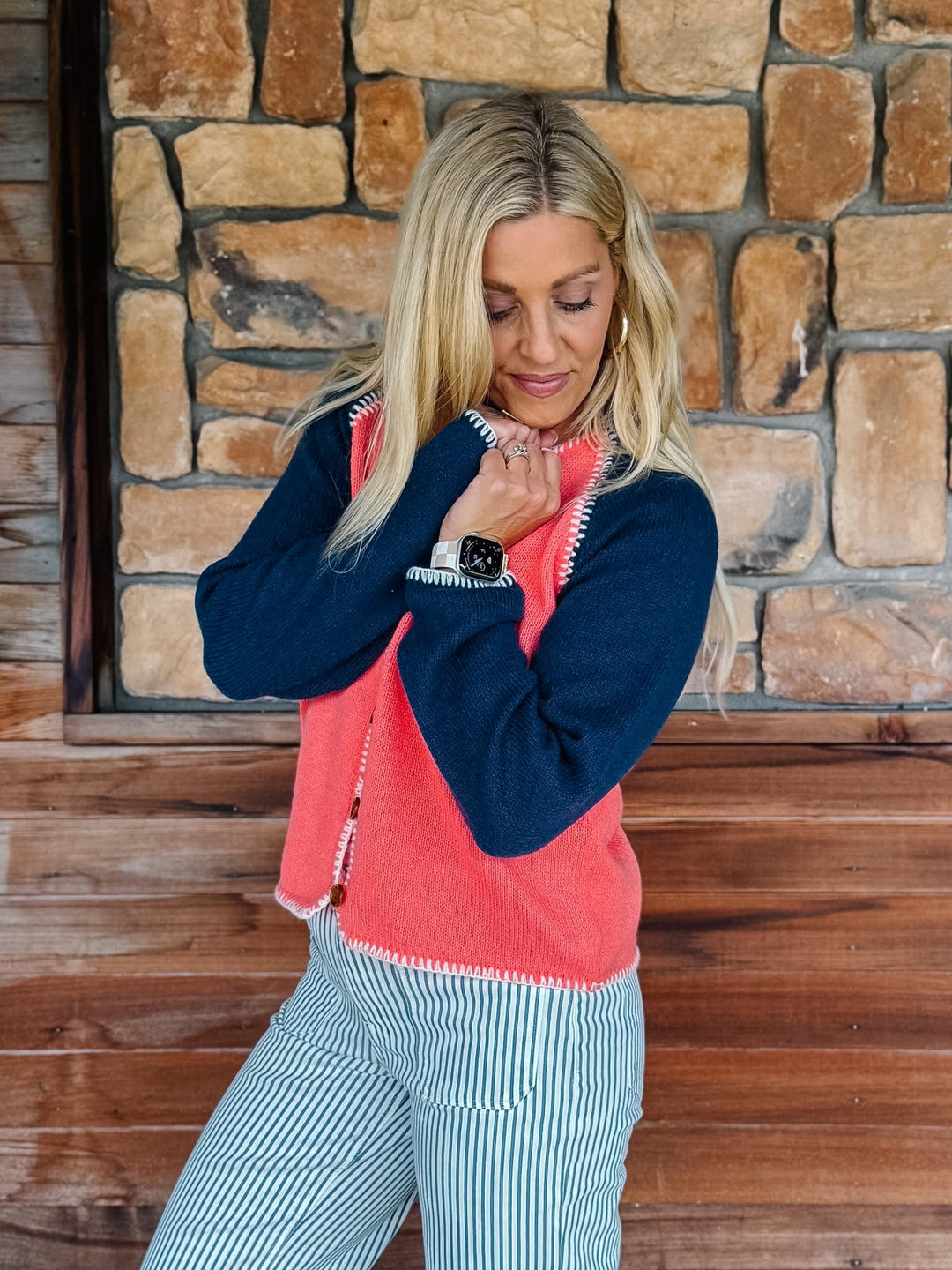 A woman wearing a coral and navy blue knit sweater vest with white zigzag trim, paired with blue and white striped pants, posing in front of a rustic stone wall.