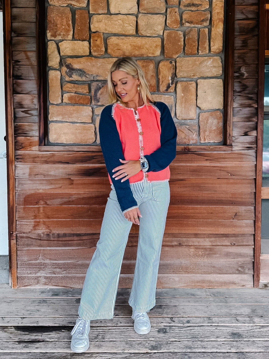 A woman stands on a rustic wooden porch in front of a stone wall, wearing a coral and navy long-sleeve top with striped light blue jeans and white sneakers.