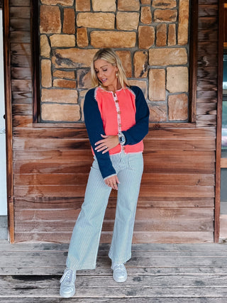 A woman stands on a rustic wooden porch in front of a stone wall, wearing a coral and navy long-sleeve top with striped light blue jeans and white sneakers.