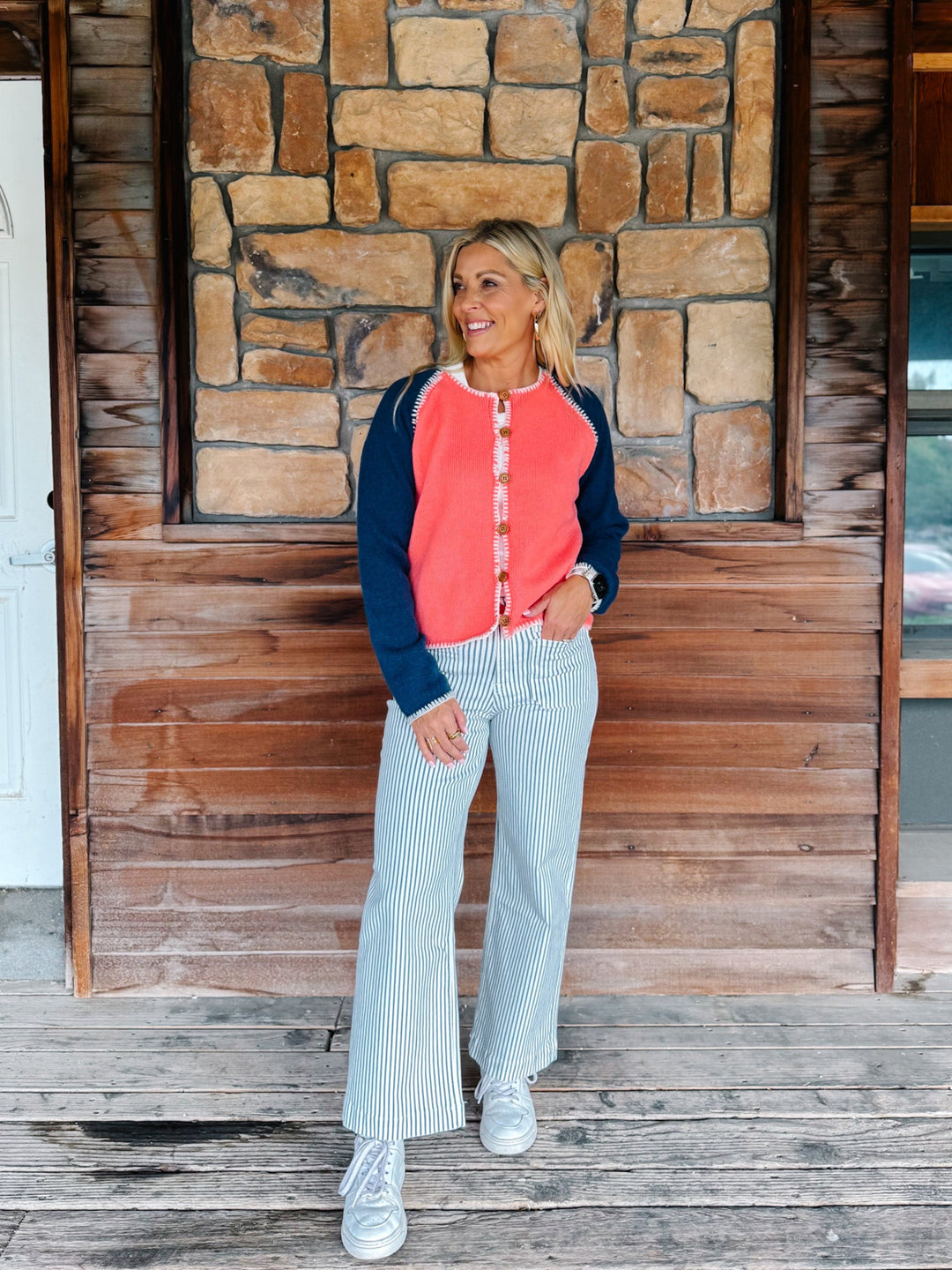 A woman stands on a wooden porch in front of a stone and wood wall, wearing a coral and navy raglan button-front top, light blue and white vertically striped pants, and white sneakers.