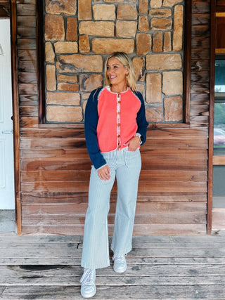 A woman stands on a wooden porch in front of a stone and wood wall, wearing a coral and navy raglan button-front top, light blue and white vertically striped pants, and white sneakers.