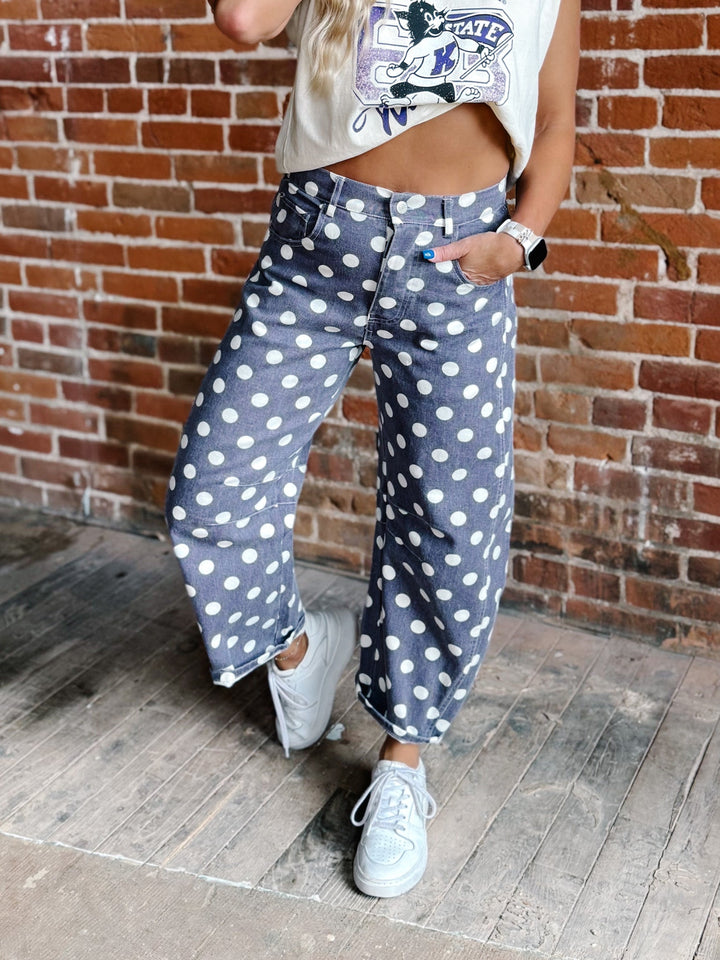A person wearing loose, wide-leg purple with a white polka dot pattern jeans, styled with a graphic crop top and white sneakers, standing against a brick wall.