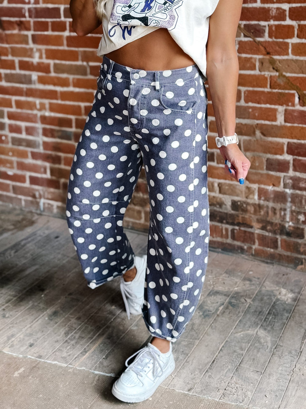 A person wearing loose, wide-leg pants with purple and white polka dots, paired with white sneakers, standing against a brick wall.
