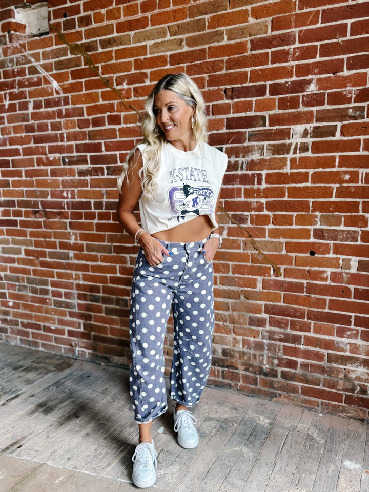 A woman stands in front of a red brick wall wearing a cream cropped graphic t-shirt, purple and white polka dot pants, and white sneakers.