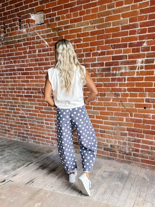 A woman with long blonde hair is shown from behind, wearing loose-fitting purple pants with white polka dots, a white sleeveless top, and white sneakers, standing in front of a red brick wall