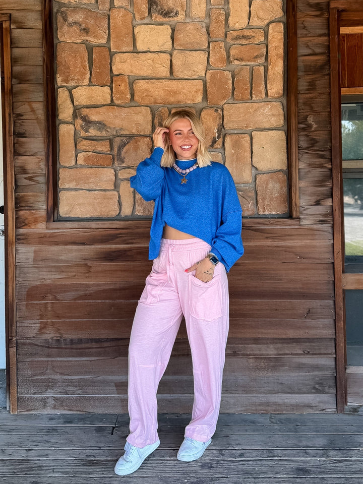 A woman stands on a wooden porch in front of a stone wall, wearing a bright blue cropped long-sleeve top, light pink wide-leg pants, and light blue sneakers.