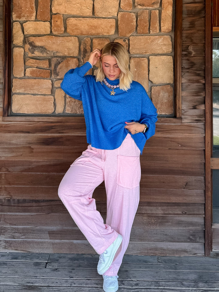 A woman posing on a rustic wooden porch in front of a stone wall, wearing a vibrant blue long-sleeve knit sweater and light pink wide-leg pants.