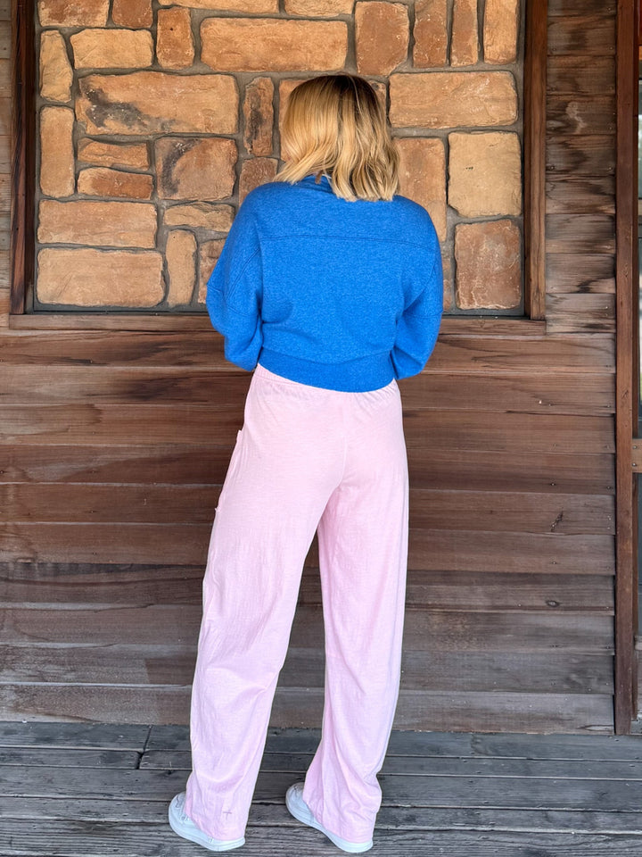 A person standing with their back to the camera, wearing a bright blue long-sleeve knit sweater and light pink wide-leg pants, positioned in front of a rustic wooden and stone wall.