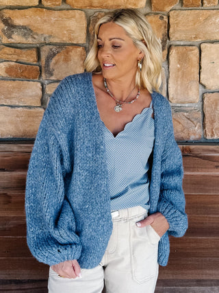 A woman wearing a chunky blue knit cardigan over a light blue scalloped top and cream pants, standing in front of a rustic stone wall.