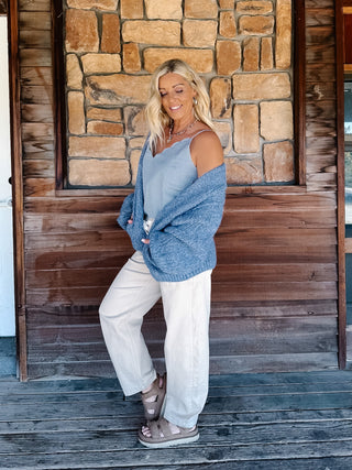 A woman wearing a chunky blue knit cardigan over a light blue scalloped top and cream pants, standing in front of a rustic stone wall.