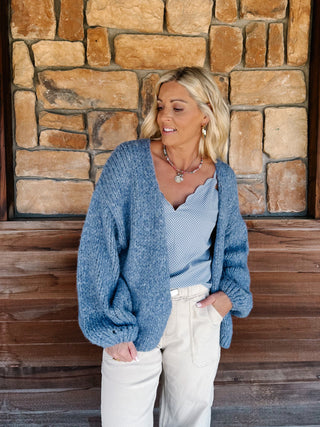 A woman wearing a chunky blue knit cardigan over a light blue scalloped top and cream pants, standing in front of a rustic stone wall.