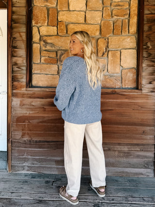 A woman wearing a chunky blue knit cardigan over a light blue scalloped top and cream pants, standing in front of a rustic stone wall.