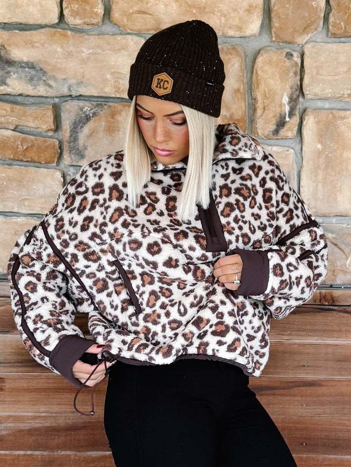 A woman wearing a brown and beige leopard print fleece pullover hoodie with dark brown trim, paired with black pants and a dark brown beanie, standing in front of a rustic stone wall.