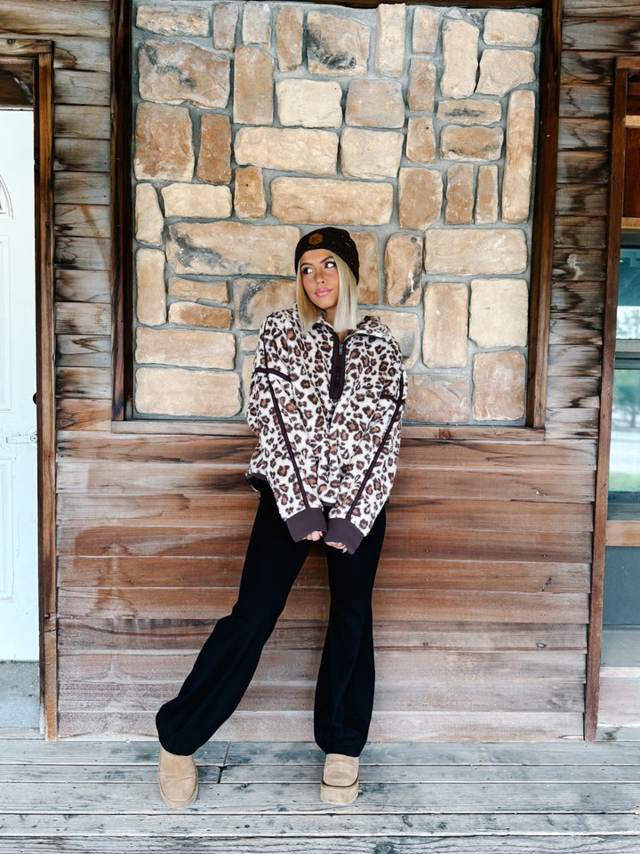 A woman wearing a brown and beige leopard print fleece pullover hoodie with dark brown trim, paired with black pants and a dark brown beanie, standing in front of a rustic stone wall.
