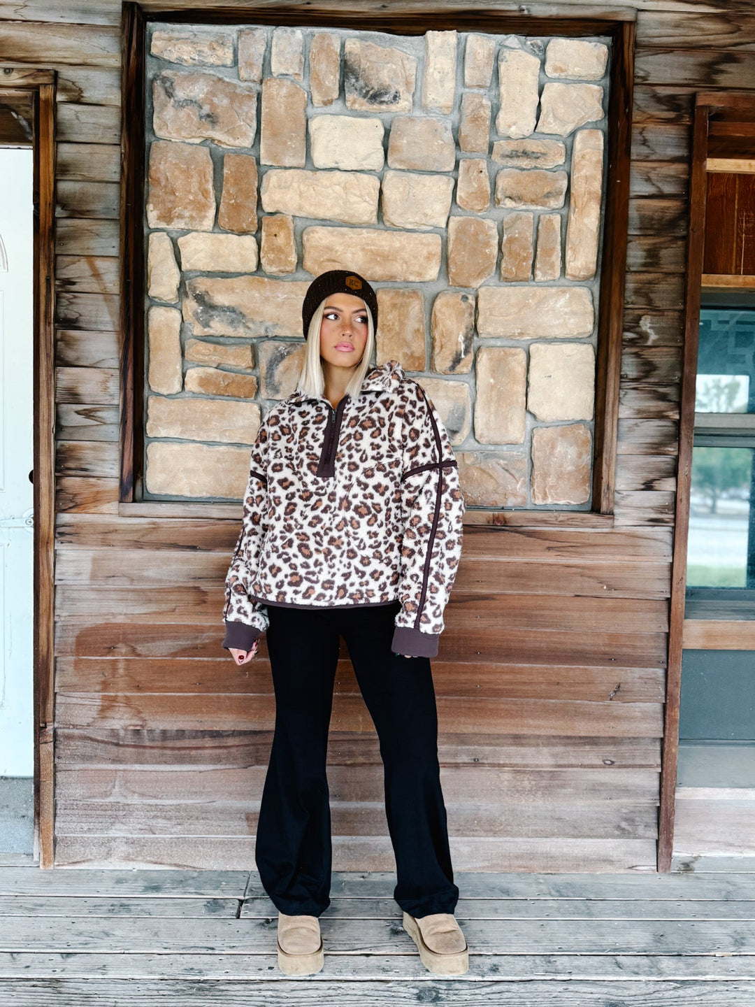 A woman wearing a brown and beige leopard print fleece pullover hoodie with dark brown trim, paired with black pants and a dark brown beanie, standing in front of a rustic stone wall.