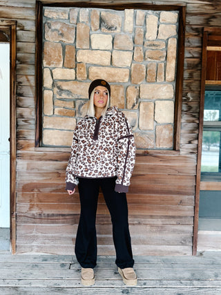 A woman wearing a brown and beige leopard print fleece pullover hoodie with dark brown trim, paired with black pants and a dark brown beanie, standing in front of a rustic stone wall.