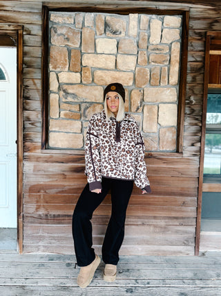 A woman wearing a brown and beige leopard print fleece pullover hoodie with dark brown trim, paired with black pants and a dark brown beanie, standing in front of a rustic stone wall.