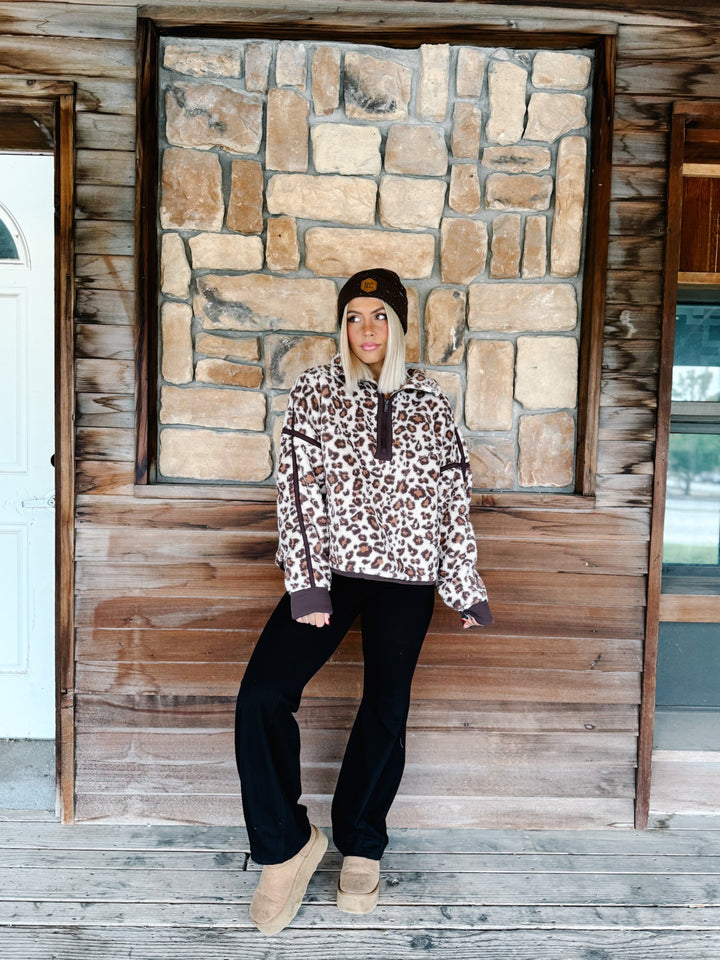 A woman wearing a brown and beige leopard print fleece pullover hoodie with dark brown trim, paired with black pants and a dark brown beanie, standing in front of a rustic stone wall.