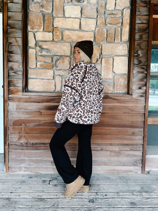 A woman wearing a brown and beige leopard print fleece pullover hoodie with dark brown trim, paired with black pants and a dark brown beanie, standing in front of a rustic stone wall.