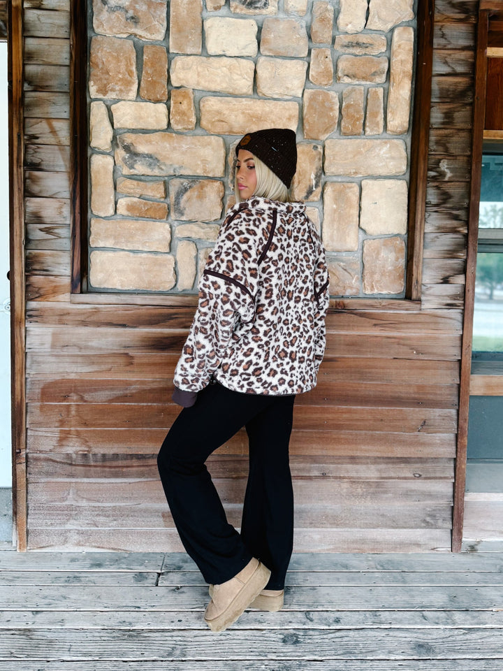 A woman wearing a brown and beige leopard print fleece pullover hoodie with dark brown trim, paired with black pants and a dark brown beanie, standing in front of a rustic stone wall.