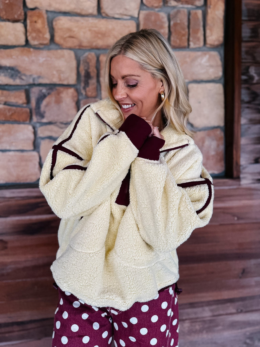 A woman wearing a cream sherpa fleece zip-up jacket with dark burgundy trim and matching burgundy cuffs, paired with burgundy polka-dot pants, standing in front of a rustic stone wall.
