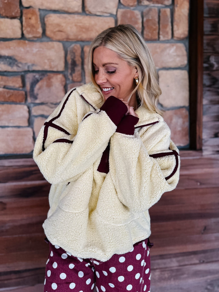A woman wearing a cream sherpa fleece zip-up jacket with dark burgundy trim and matching burgundy cuffs, paired with burgundy polka-dot pants, standing in front of a rustic stone wall.