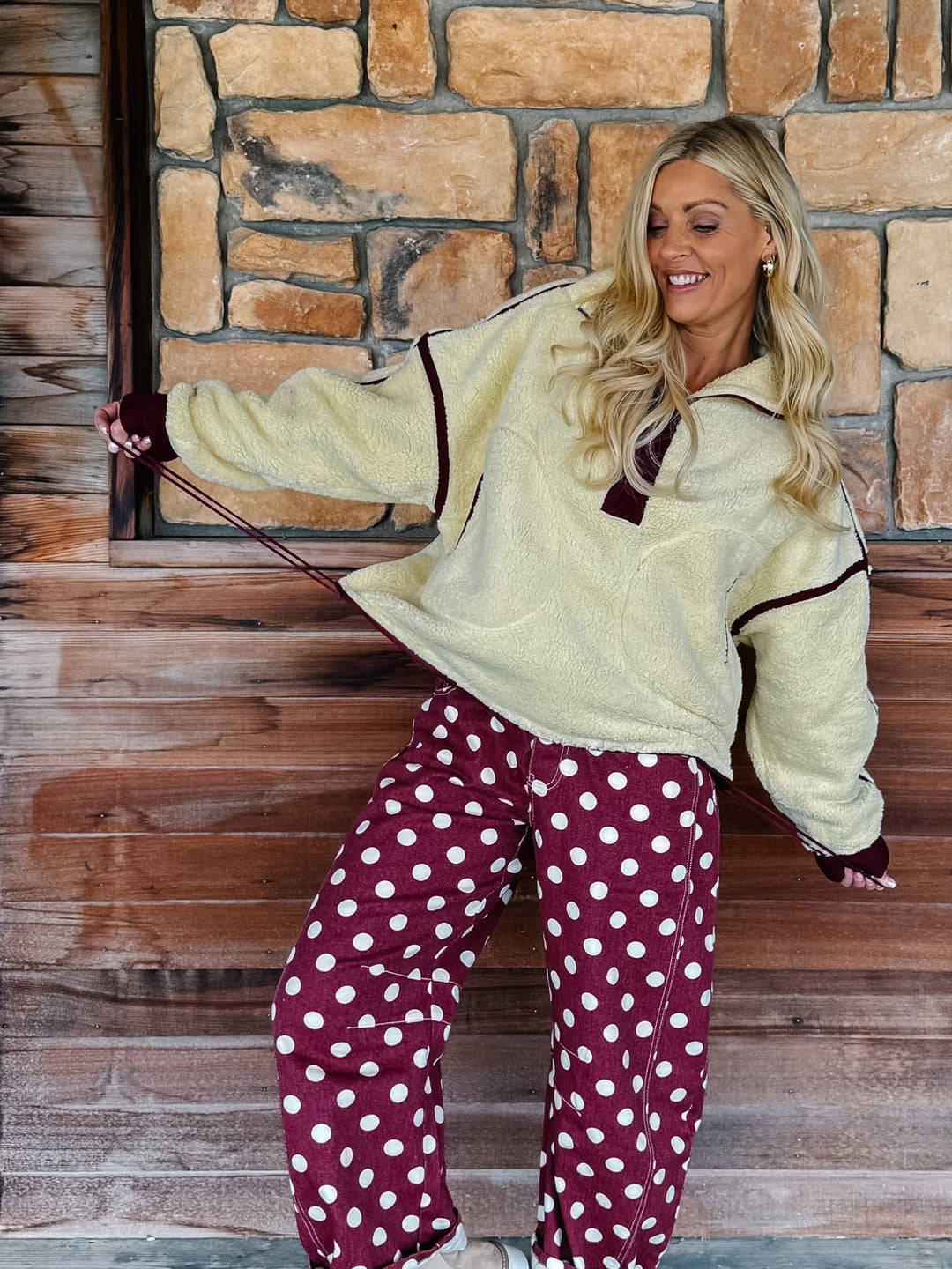 A woman wearing a cream sherpa fleece zip-up jacket with dark burgundy trim and matching burgundy cuffs, paired with burgundy polka-dot pants, standing in front of a rustic stone wall.