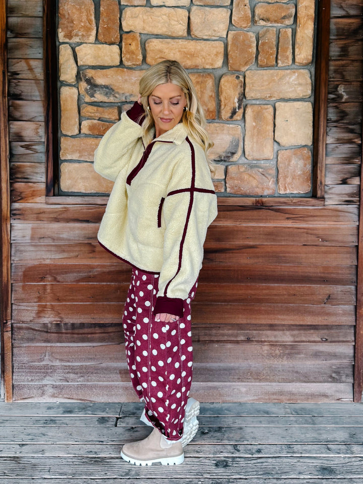 A woman wearing a cream sherpa fleece zip-up jacket with dark burgundy trim and matching burgundy cuffs, paired with burgundy polka-dot pants, standing in front of a rustic stone wall.