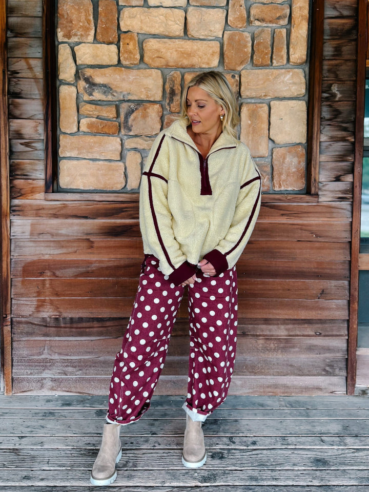 A woman wearing a cream sherpa fleece zip-up jacket with dark burgundy trim and matching burgundy cuffs, paired with burgundy polka-dot pants, standing in front of a rustic stone wall.