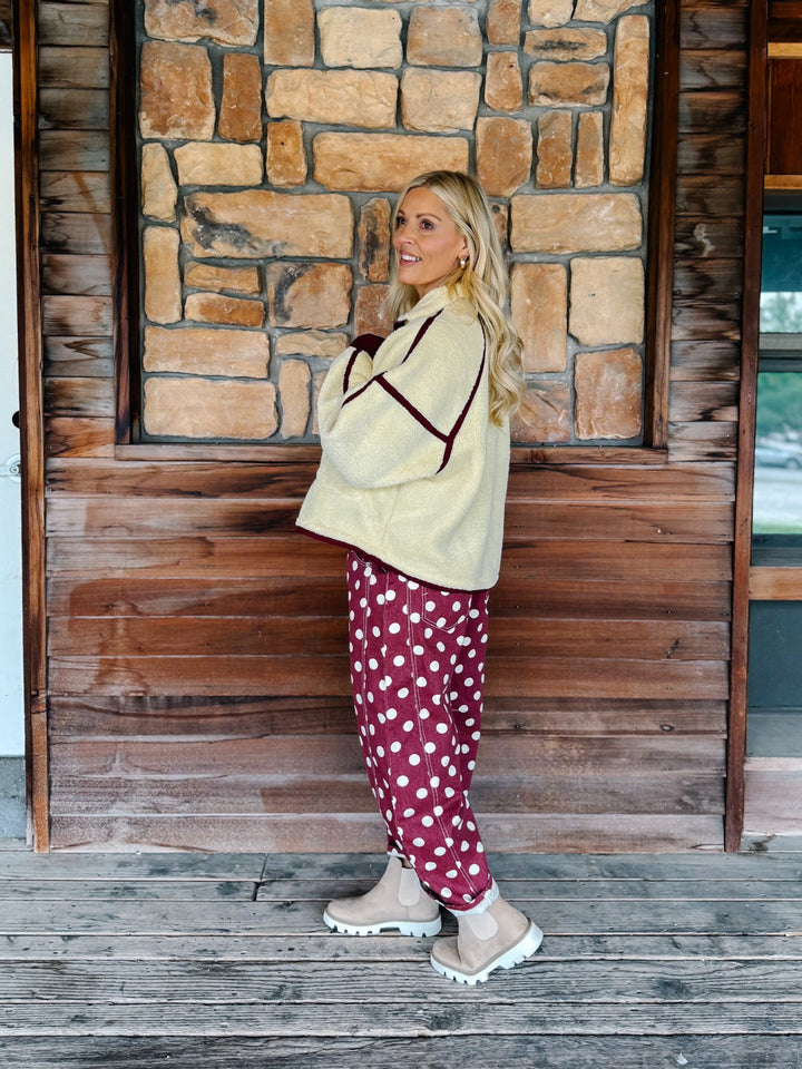A woman wearing a cream sherpa fleece zip-up jacket with dark burgundy trim and matching burgundy cuffs, paired with burgundy polka-dot pants, standing in front of a rustic stone wall.